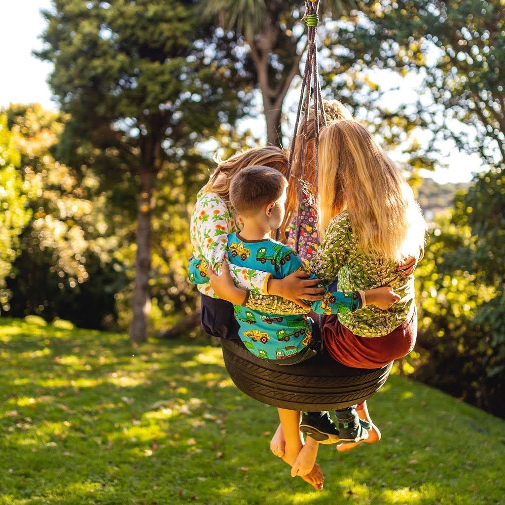 Four kids swing on a tire over a grassy backyard, dressed in colourful clothes from Hoopla Kids, and in bare feet. The late afternoon sunlight is dappled by trees..