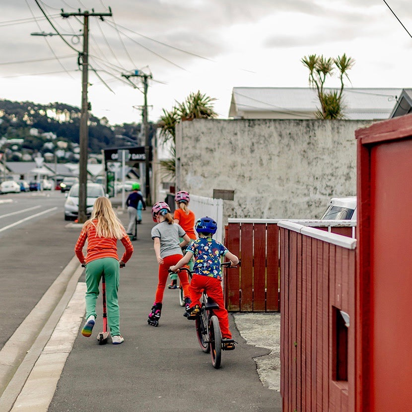 Hoopla Kids riding their wheels down a Rongotai street one grey afternoon, wearing bright coloured clothes from the Hoopla Kids Stockroom